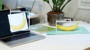 A banana on a table and an image of a banana on a laptop on the same table. Each of the two bananas has a white frame around it with the word ‚Banana‘ sticked on top of it.