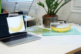 A banana on a table and an image of a banana on a laptop on the same table. Each of the two bananas has a white frame around it with the word ‚Banana‘ sticked on top of it.