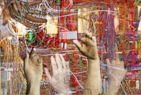 The image depicts a series of stone-like hands, fractured and cracked, reaching out in various directions from a chaotic mass of tangled wires. Some hands display broken fingers, while archival tags on others suggest anonymity and erasure. The juxtaposition of human forms and technological wires evokes themes of labor, exploitation, and the often unseen physical work behind the tech industry.