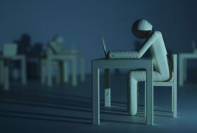Plastic figure resembling a human who sits on a table infront of a laptop in a dark room. Long shadows disseminate a gloomy mood.