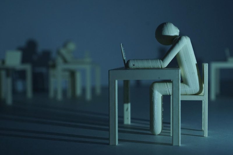 Plastic figure resembling a human who sits on a table infront of a laptop in a dark room. Long shadows disseminate a gloomy mood.